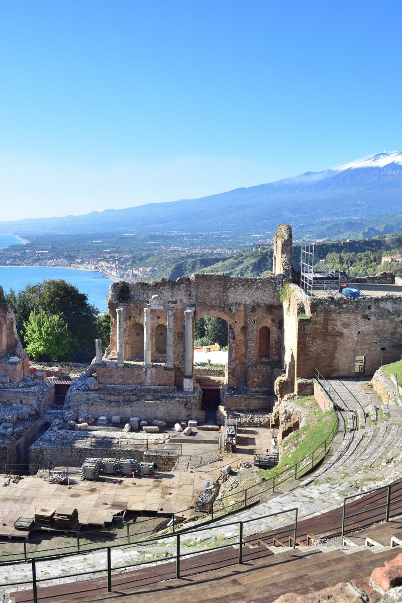 The rock-cut cavea of the Ancient Theatre of Taormina, with Mount Etna and the Ionian Sea framed beyond the ruined stage wall